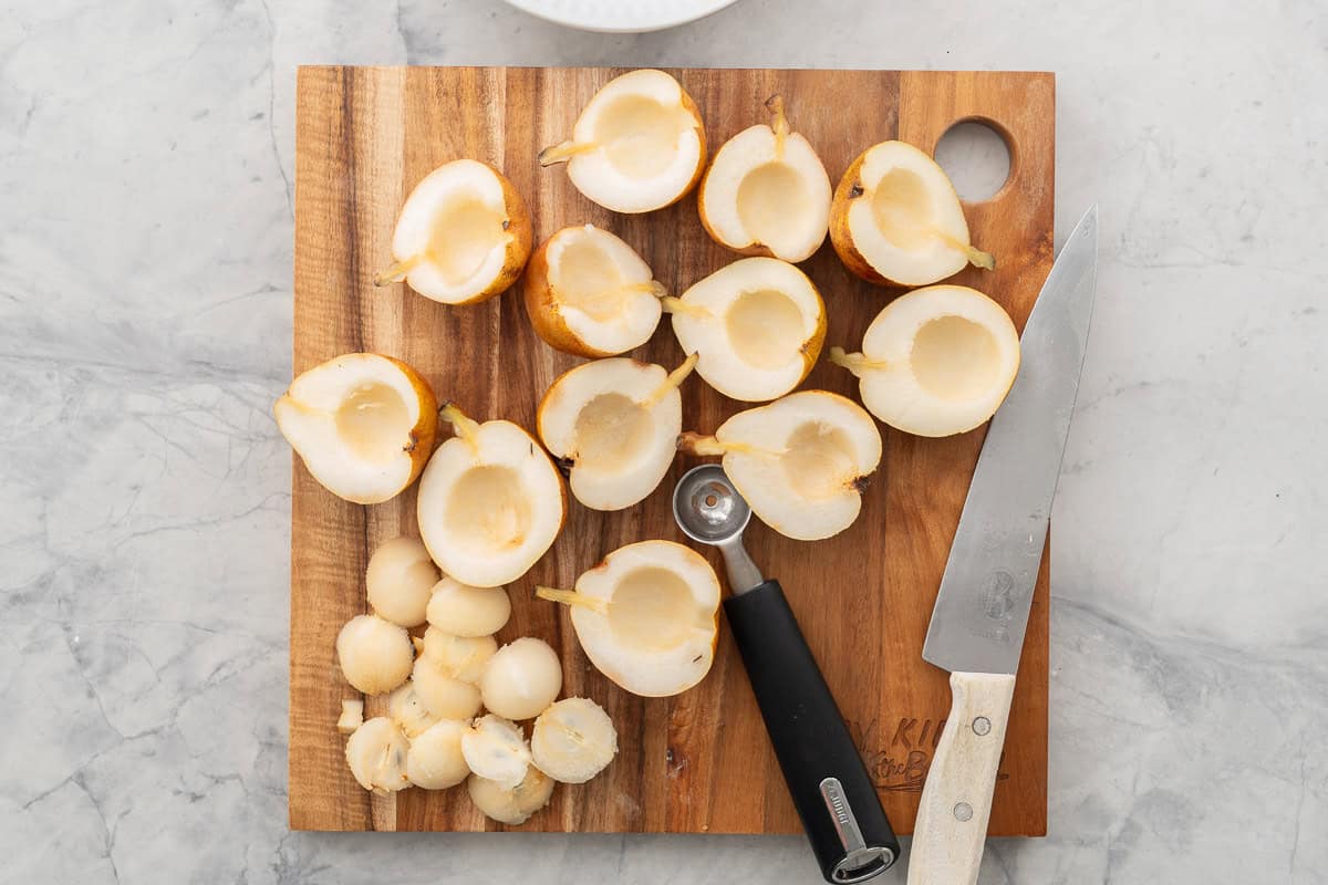 Fresh pears cut in half with cores scooped out of the middle sitting on a wooden chopping board with knife and melon baller.