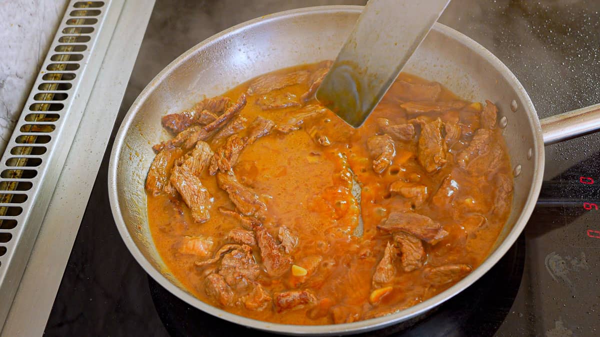 Strips of beef in a glossy sauce that is boiling in a stainless steel skillet. 