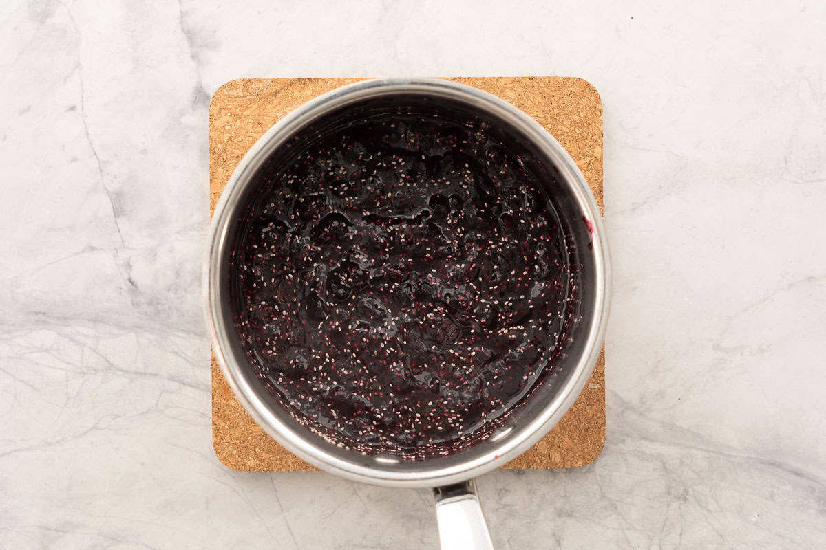 Blueberry chia jam in pot on wooden place mat on bench top.