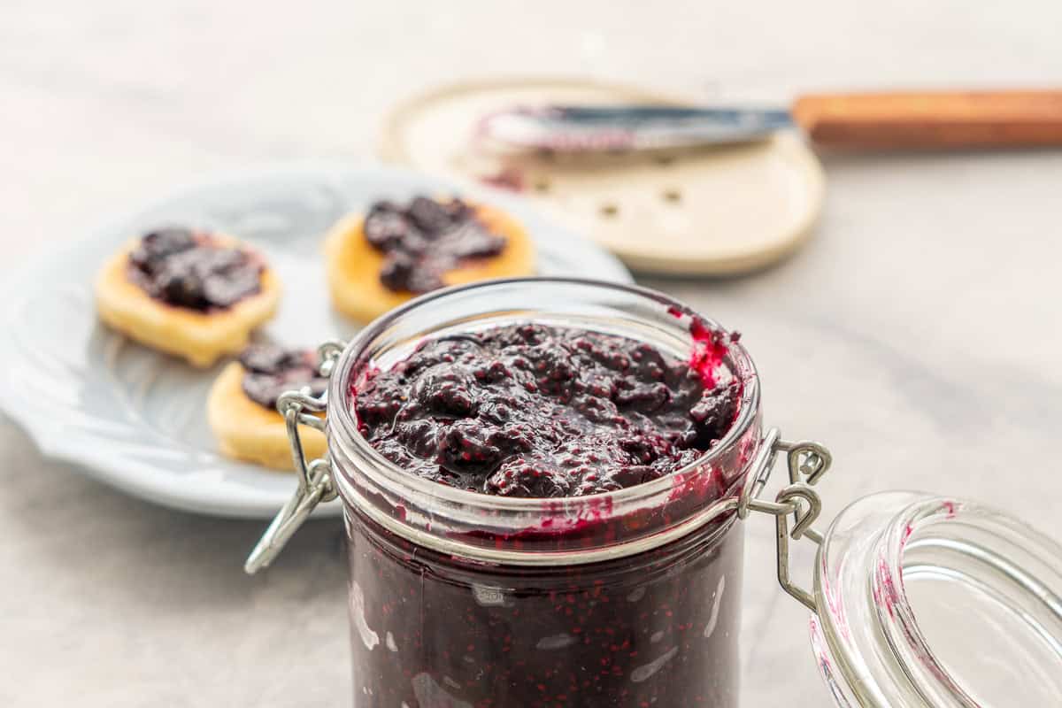 Chia Blueberry jam in jar with lid open filled to the top, in background a plate mini pancakes with jam spread on them.