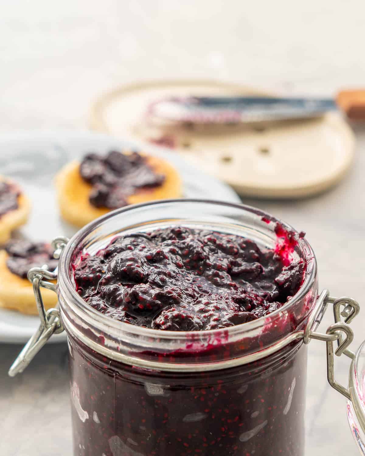 Chia Blueberry jam in jar with lid open filled to the top, in background a plate mini pancakes with jam spread on them.