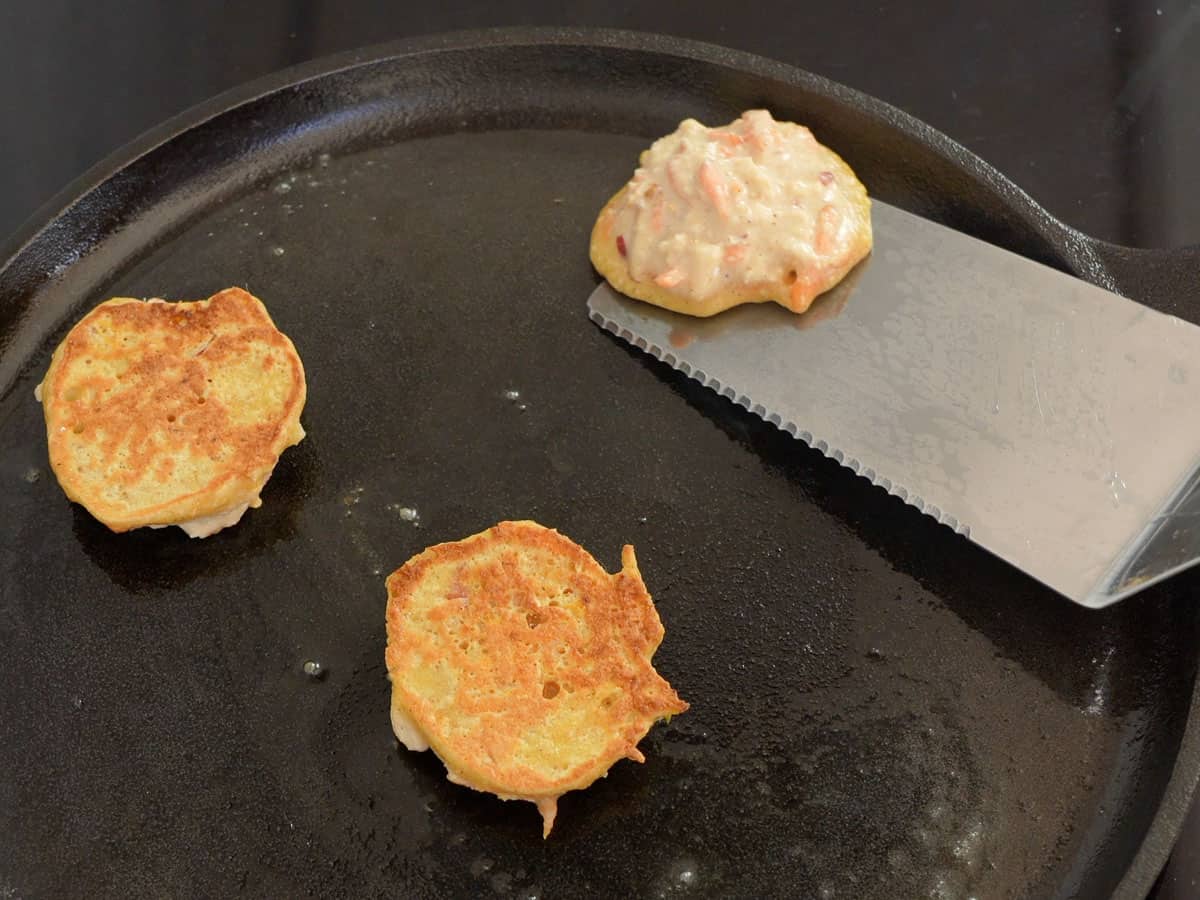 Carrot cake pancakes in pan two cooked and one being flipped with metal spatular showing uncooked side.