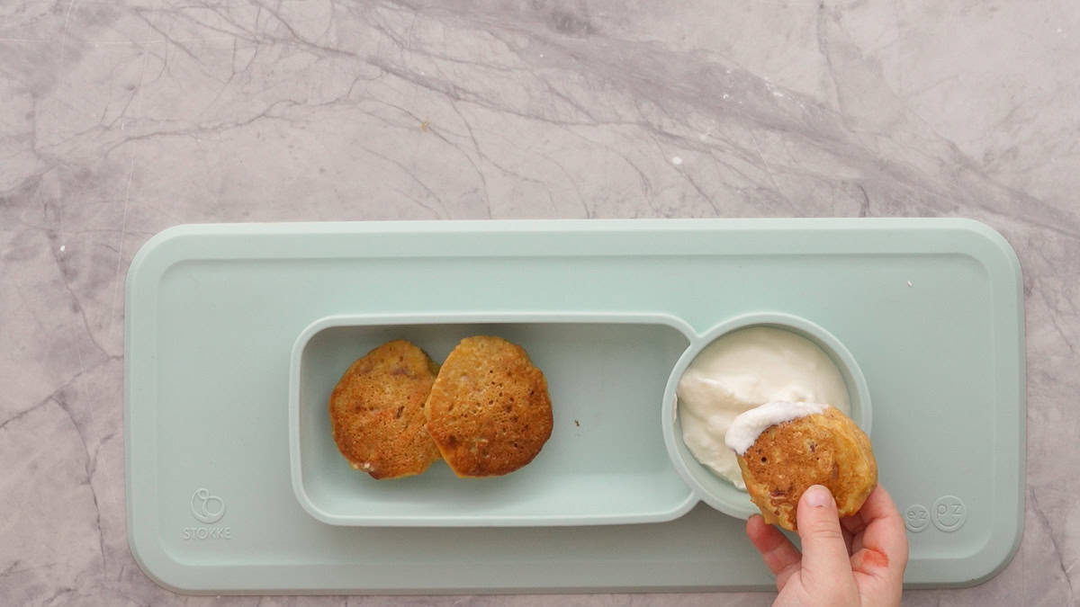 Three carrot cake pancakes in silicone baby plate with greek yogurt, one pancake being dipped into the yogurt by a toddlers hand.