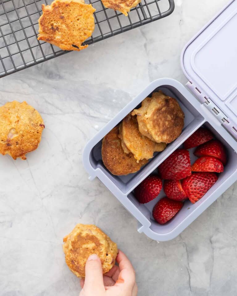 Childs lunch box with divider through the centre with carrot cake pancakes on one side and halved strawberries. Child's hand holding a pancake.