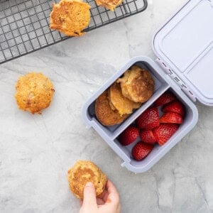 Childs lunch box with divider through the centre with carrot cake pancakes on one side and halved strawberries. Child's hand holding a pancake.