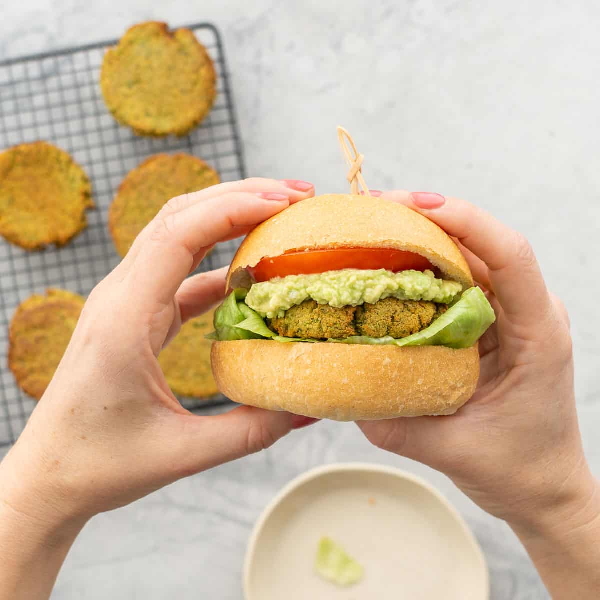 Hands holding up a Burger with lettuce, chickpea patty, guacamole, slice of tomato and top of burger.