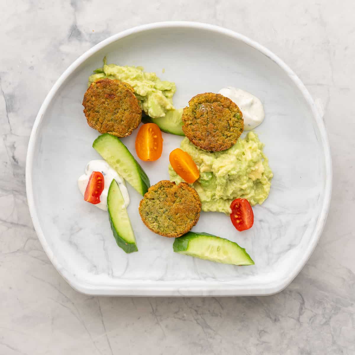 A childs silicone plate with three mini chickpea patties, aioli, guacamole, slices of cherry tomatoes and cucumber.