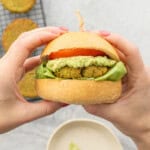 Hands holding up a Burger with lettuce, chickpea patty, guacamole, slice of tomato and top of burger.