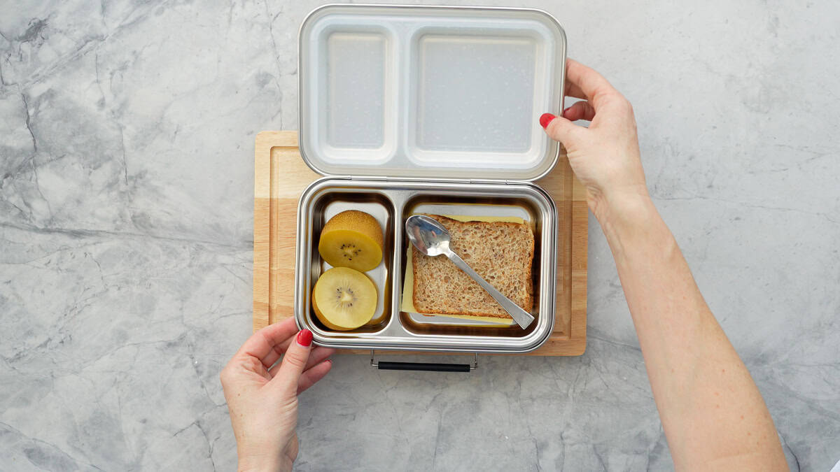 Hands opening the lid of a lunchbox with Cut Kiwifruit in one section and a sandwich with teaspoon laid on top on other section.