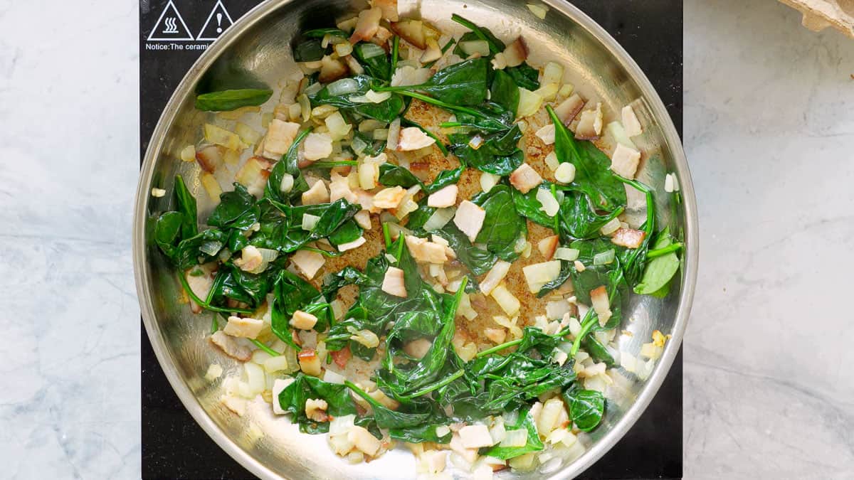 A stainless steal skillet of bacons, onions and wilted spinach. 