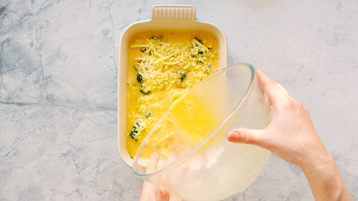 Whisked eggs in a large glass mixing bowl being poured over a baking dish filled with hash browns and spinach. 