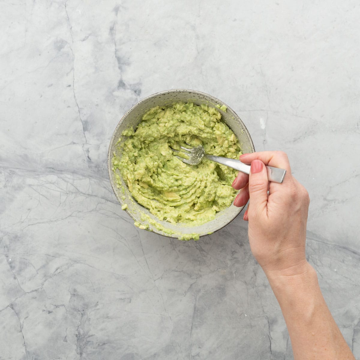 Avocado being mashed with a fork in bowl combining with garlic, lemon juice and salt.