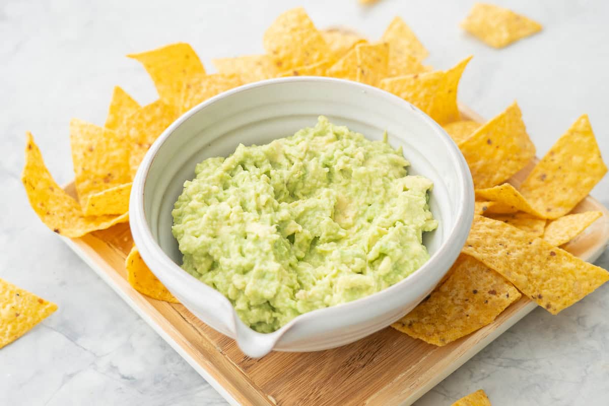 Guacamole in a bowl on wooden board surrounded by nacho chips.