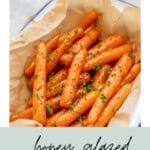 Carrots honey glazed roasted and placed on brown baking paper in serving dish, garnished with chopped parsley with text overlay for Pinterest.