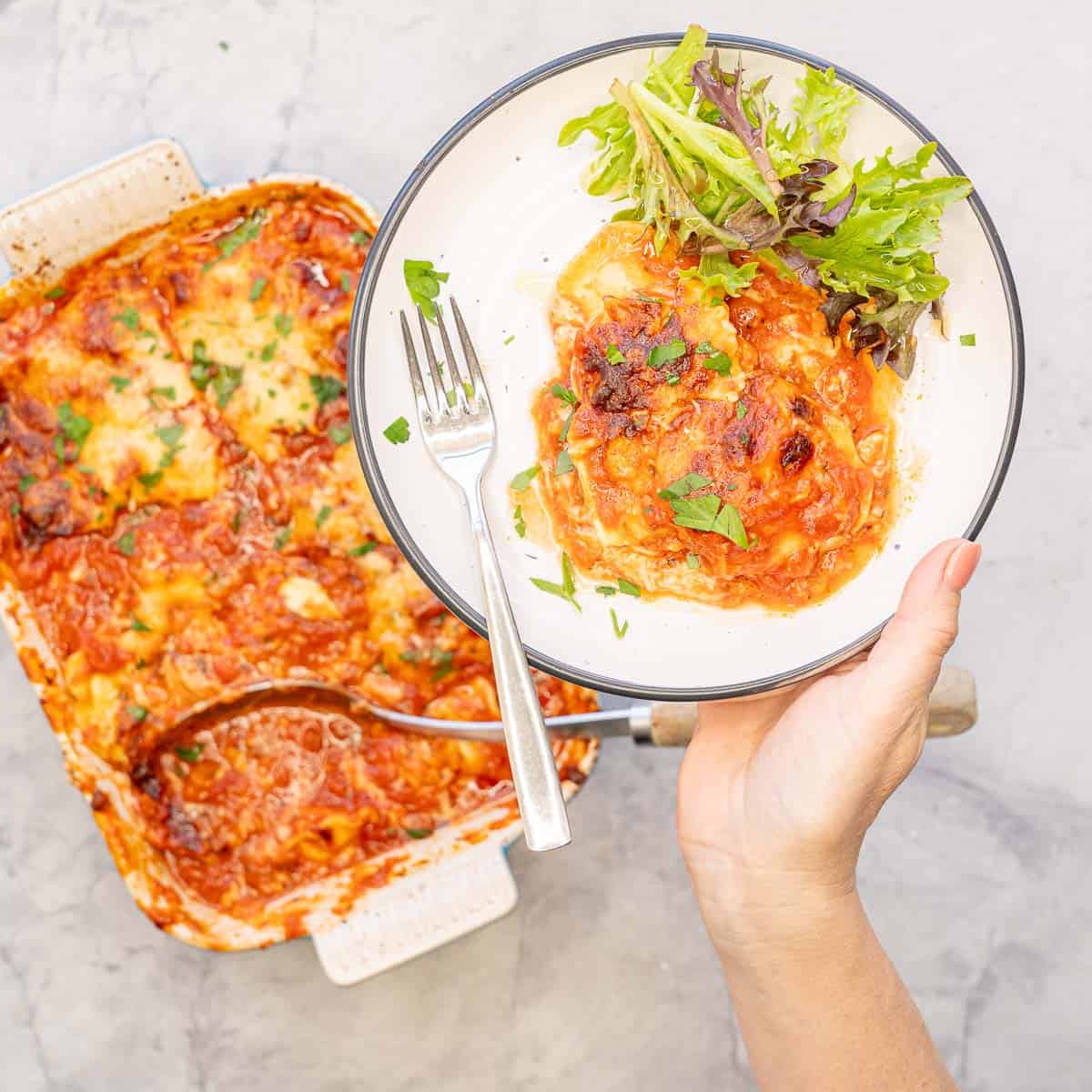 Hand holding up Lazy lasagna served on plate with side salad and fork, baking dish with serving spoon lasagna blurred in background.