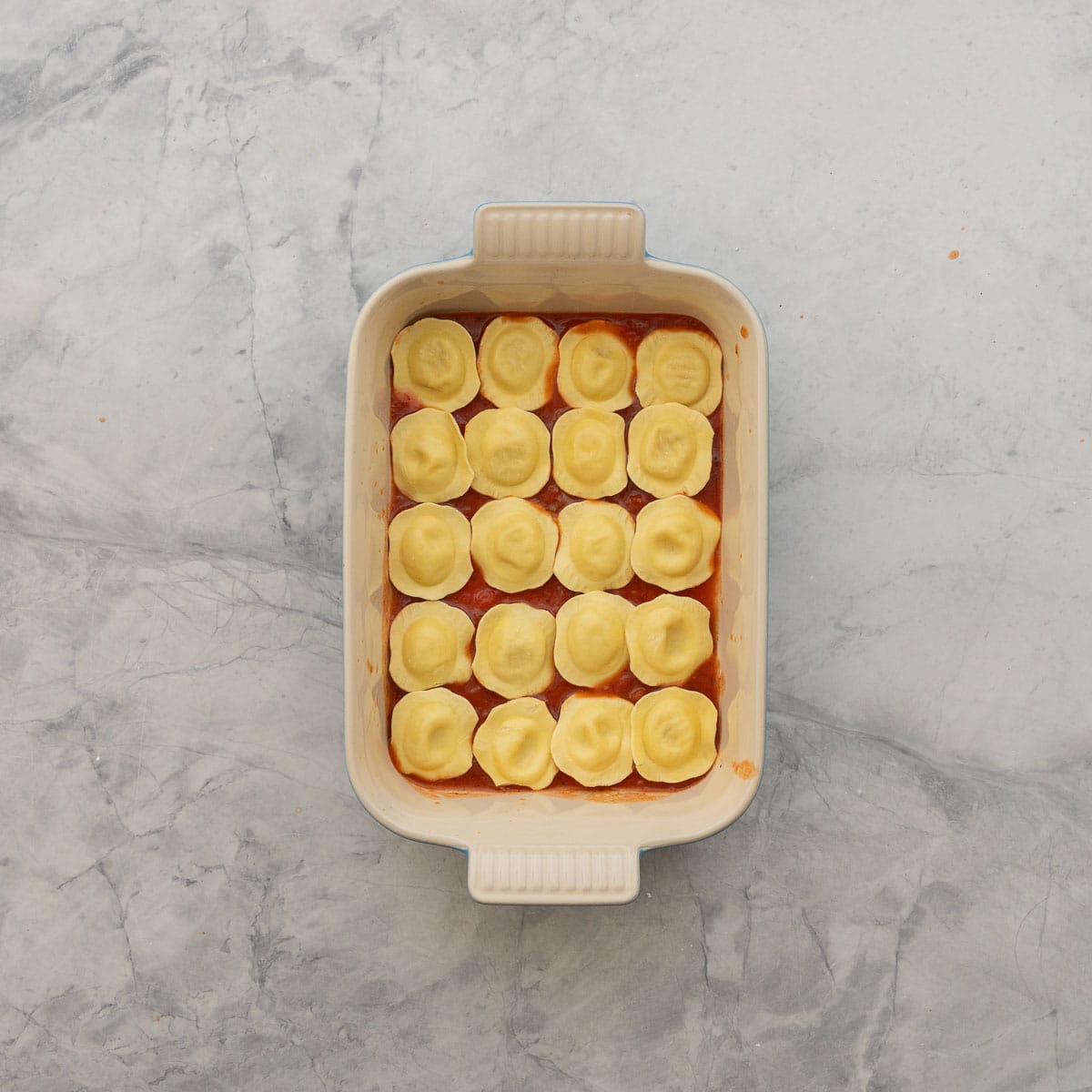 Layer of pasta sauce and ravioli in baking dish.