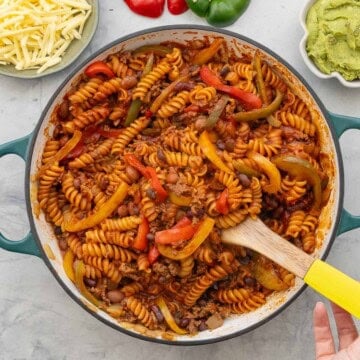 Mexican Pasta in pot with Wooden serving spoon. Grated cheese, Capsicum and guacamole on bench top in bowls.