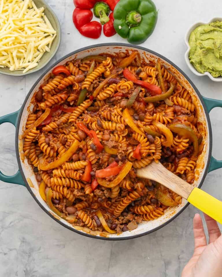 Mexican Pasta in pot with Wooden serving spoon. Grated cheese, Capsicum and guacamole on bench top in bowls.