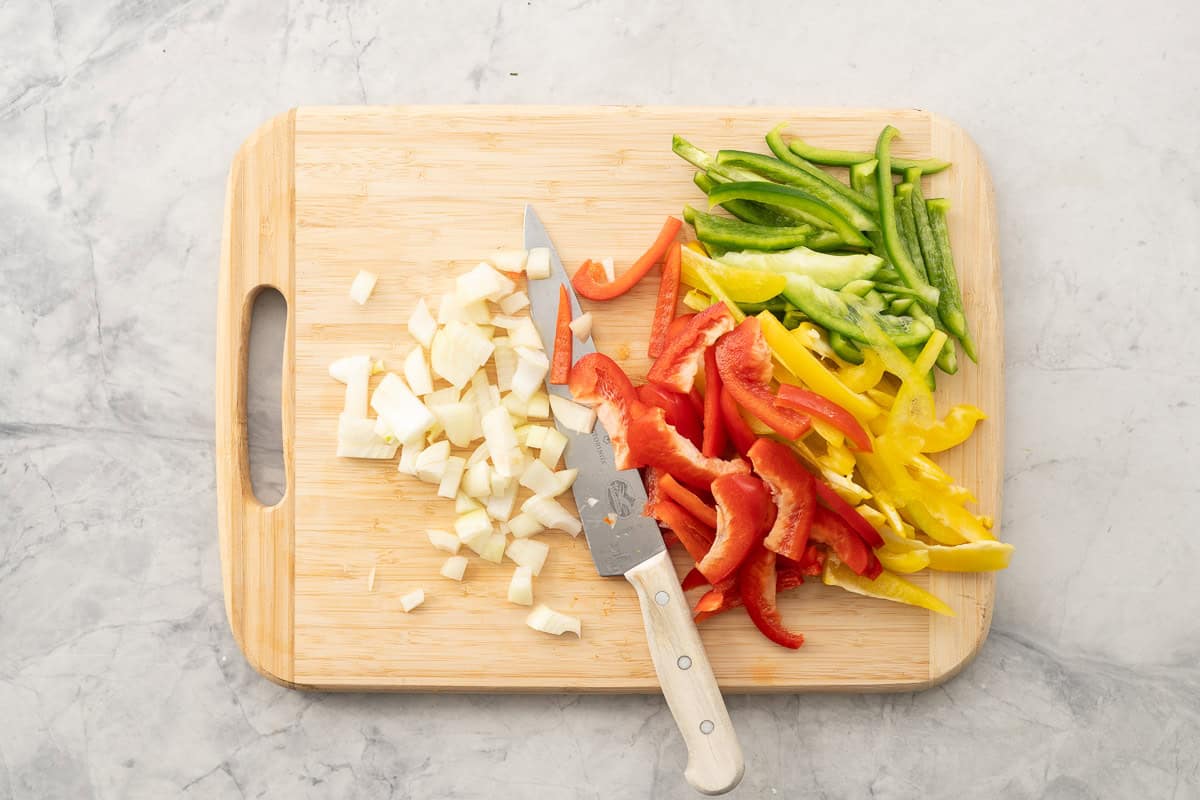 Chopped onion, sliced red, yellow and green capsicum on chopping board with knife.