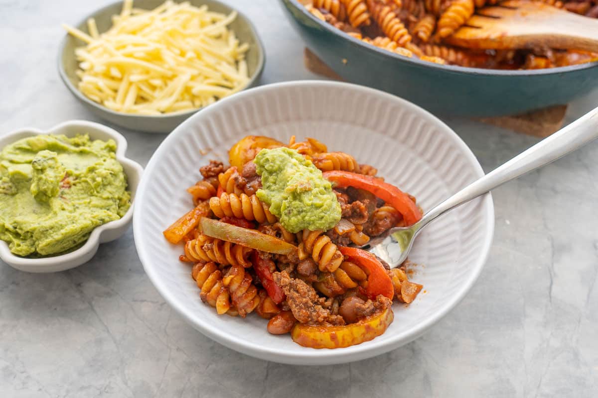 Mexican Pasta in dinner plate with fork and guacamole on top. Grated cheese, and guacamole on bench top in bowls.