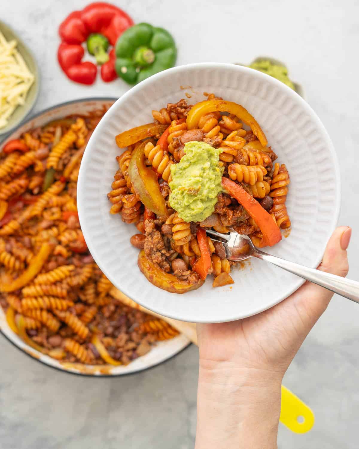 Hand holding up Mexican Pasta in dinner plate with fork and guacamole on top. Grated cheese, capsicum and guacamole on bench top in bowls.