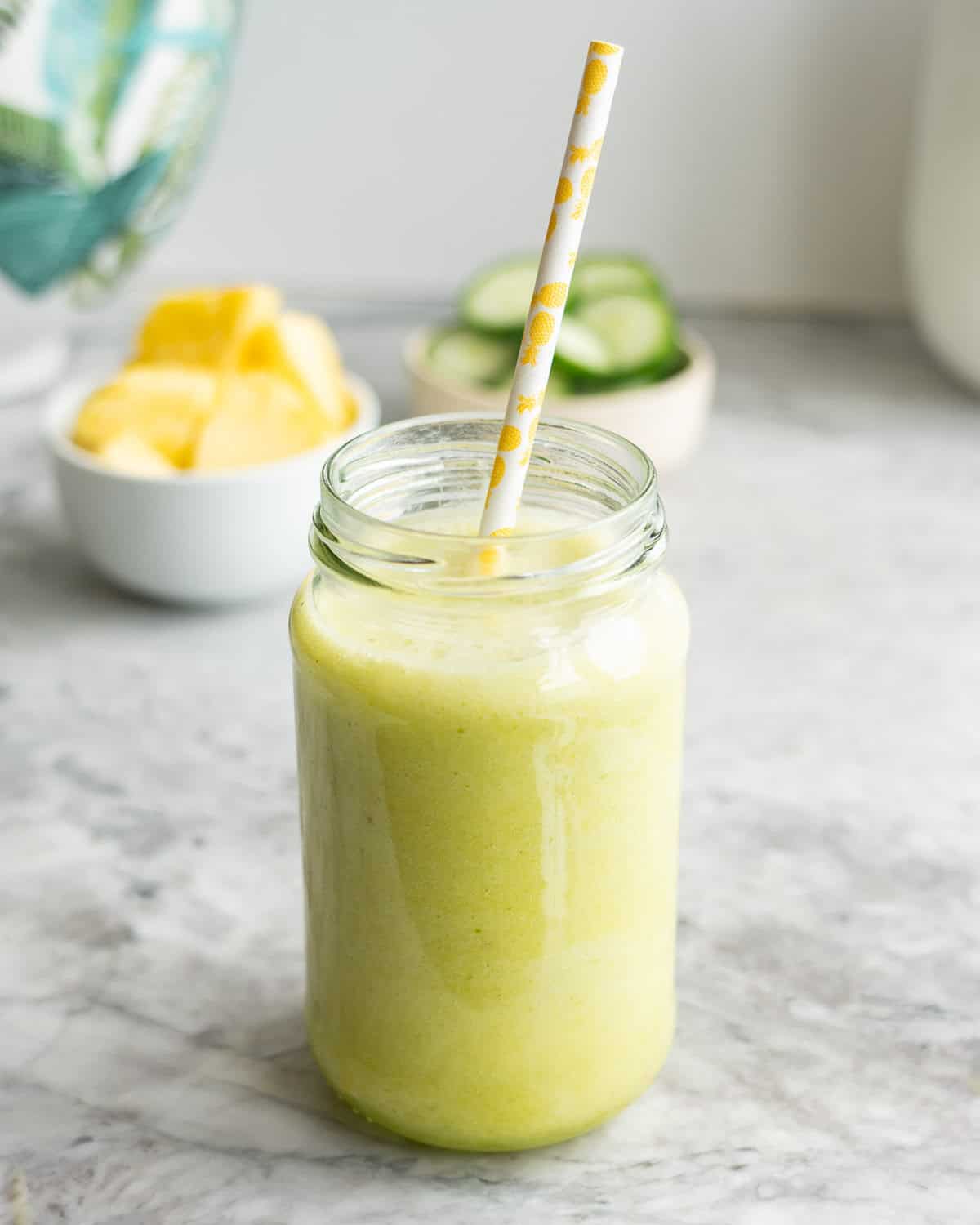 Pineapple cucumber smoothie in tall glass jar with straw. Chopped pineapple and cucumber blurred in background.