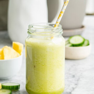 Pineapple cucumber smoothie in tall glass jar with straw. Chopped pineapple and cucumber blurred in background.