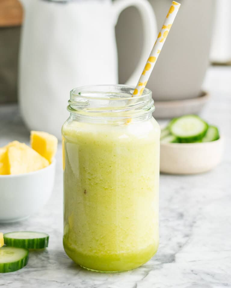 Pineapple cucumber smoothie in tall glass jar with straw. Chopped pineapple and cucumber blurred in background.