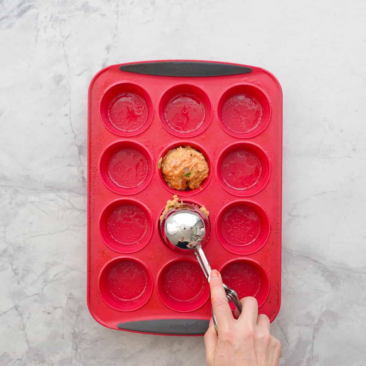 Pizza muffin batter being scooped with a cookie scooper, going into greased silicone muffin tray.