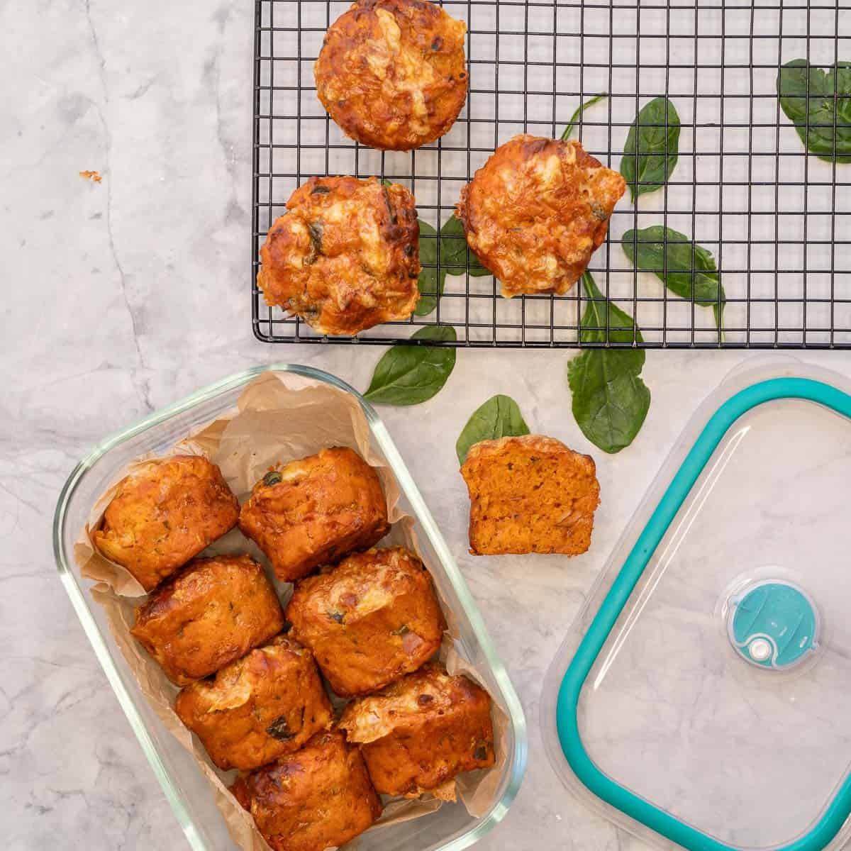 Pizza muffins in glass storage container with lid on bench top, cooling rack with three muffins on and some loose spinach leaves on bench top.