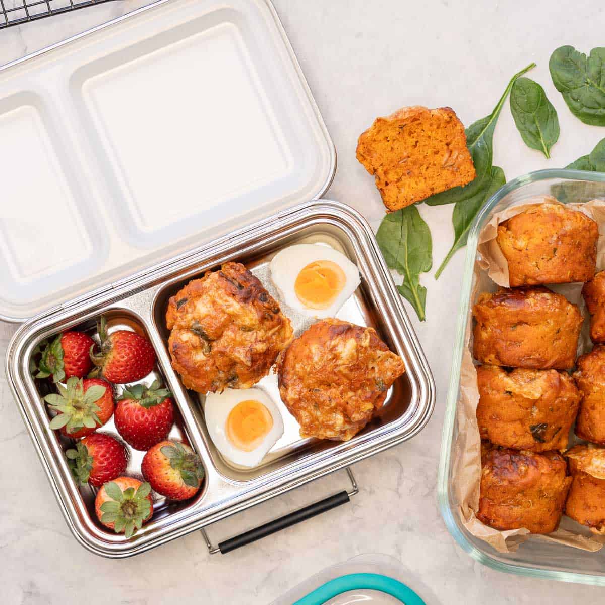 Pizza muffins in glass storage container along with lunchbox with two Pizza muffins, a cut open hardball egg and fresh strawberries.