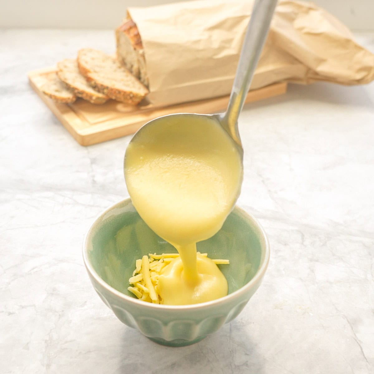 Potato soup being ladled into a bowl on top of some grated cheese. Sliced fresh bread in background on chopping board.