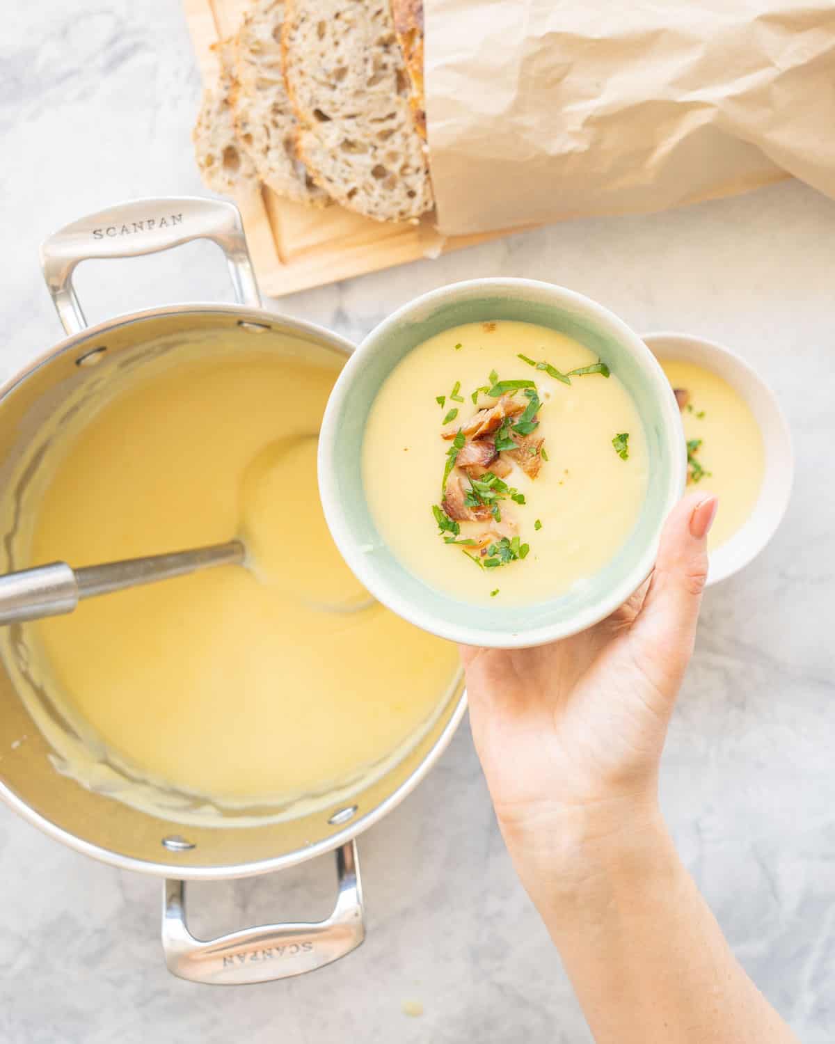 Hand holding a bowl of potato soup garnished, held above the fill pot of soup with ladle inside.