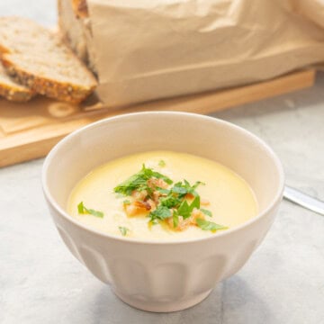 Potato soup garnished and served in bowl. Sliced fresh bread on chopping board in background.