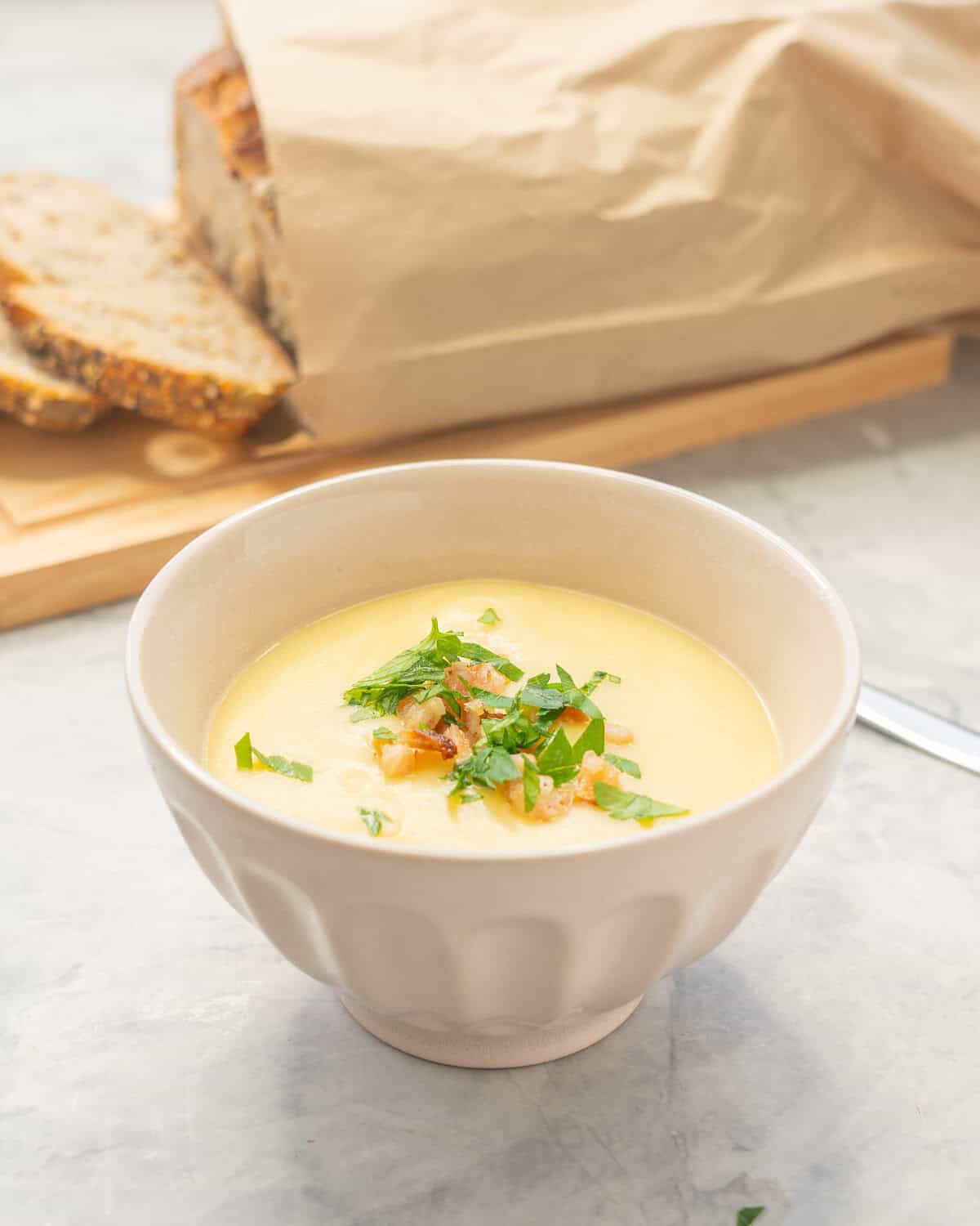 Potato soup garnished and served in bowl. Sliced fresh bread on chopping board in background.