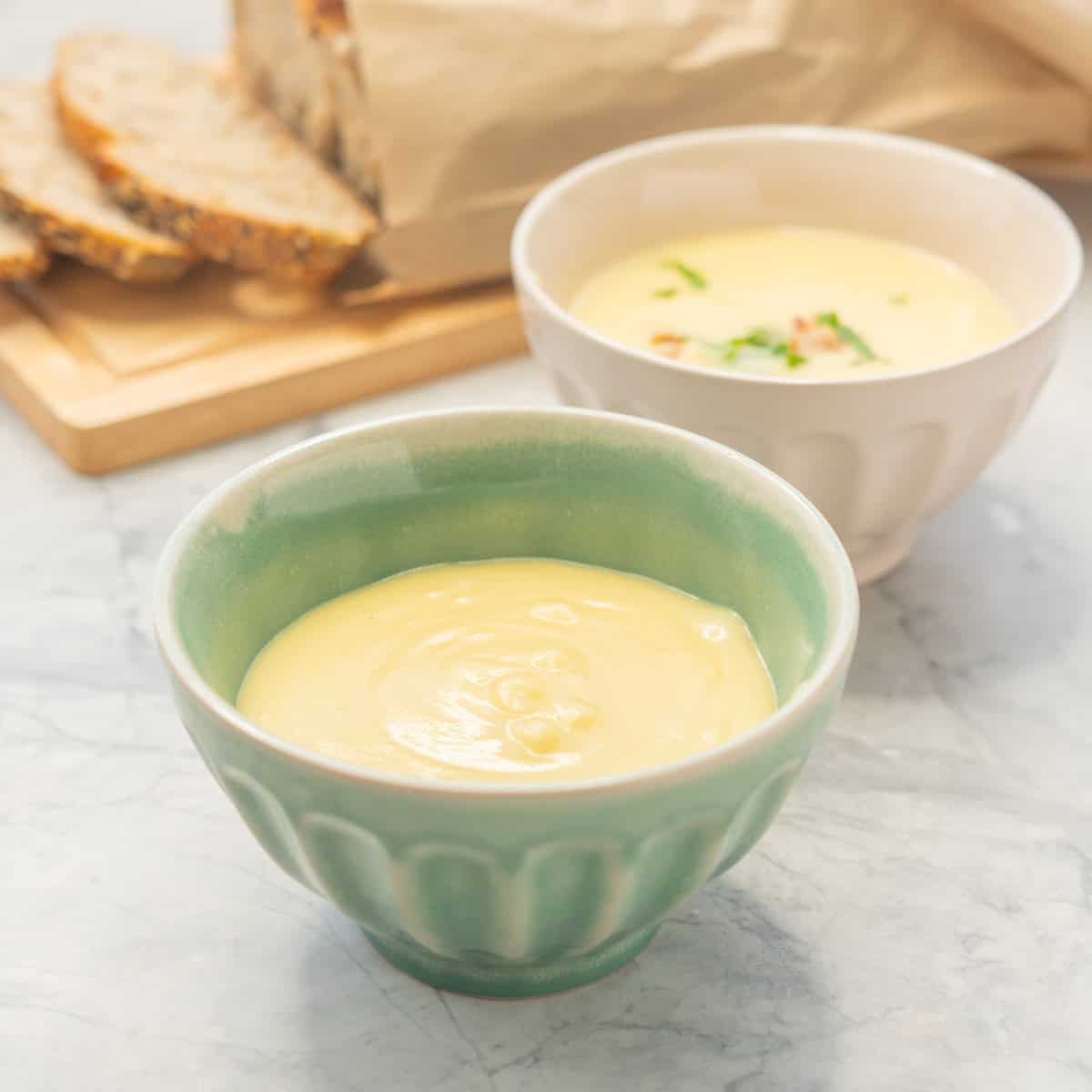 Two bowls of Potato soup one garnished. Sliced fresh bread on chopping board in background.