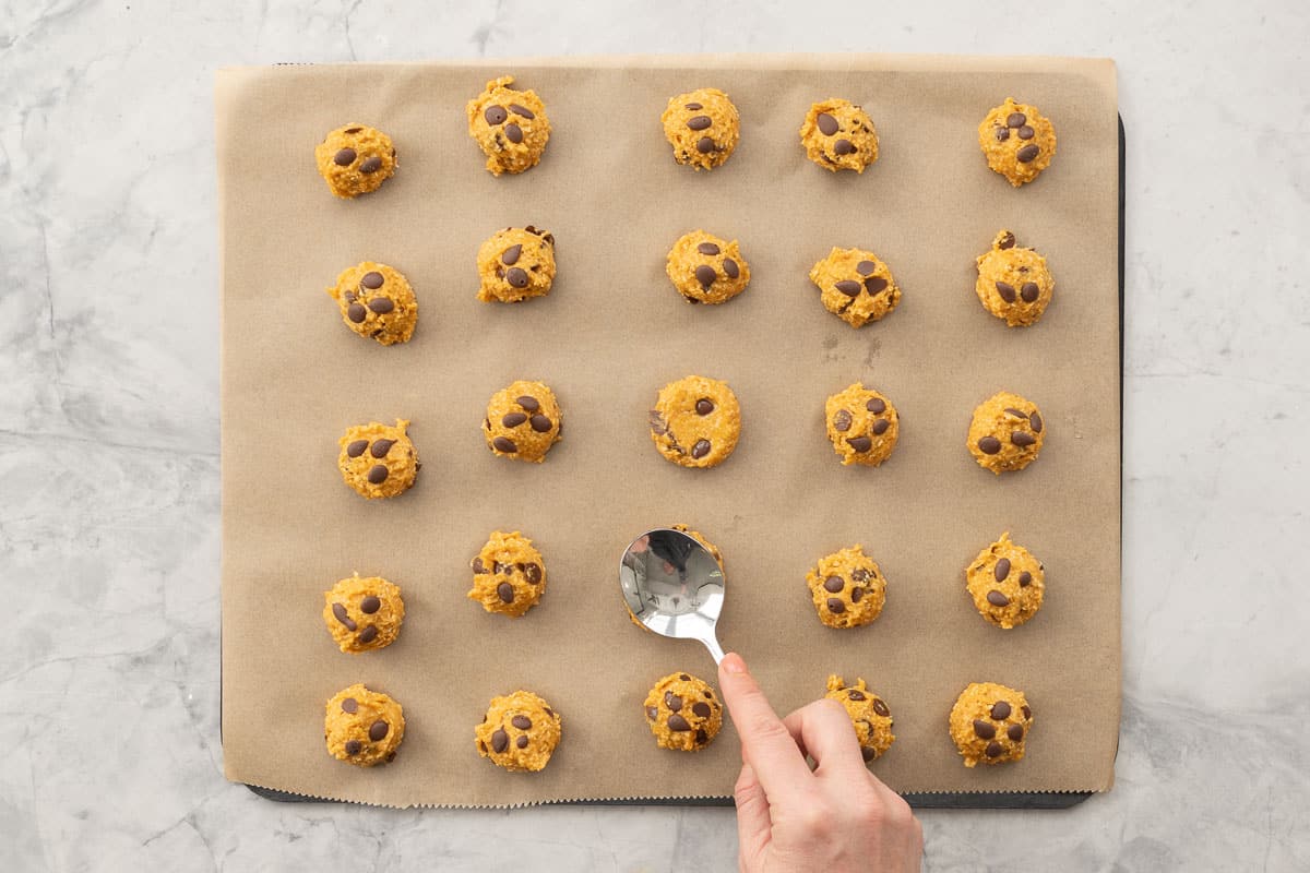 A baking tray of Pumpkin cookies rolled into balls with chocolate chips throughout, hand holding a desert spoon carefully pushing down to slightly flatten cookies.