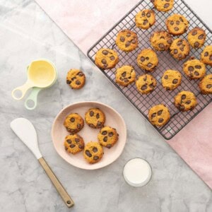 A cooling rack of Pumpkin and chocolate chip cookies, a pink ceramic plate with five cookies on it and a glass a milk. Some baking utensils on bench top.