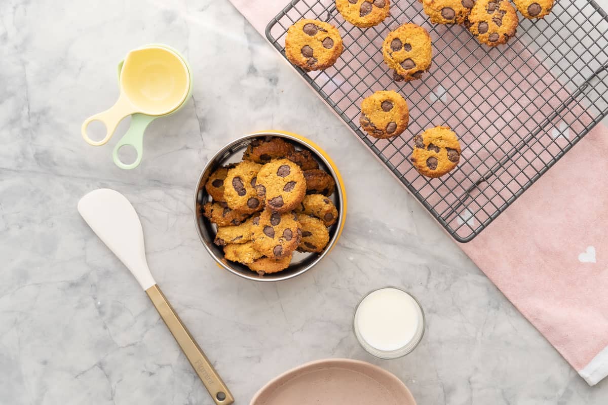 A cooling rack of Pumpkin and chocolate chip cookies and a tin with some inside. A glass a milk and baking utensils on bench top.