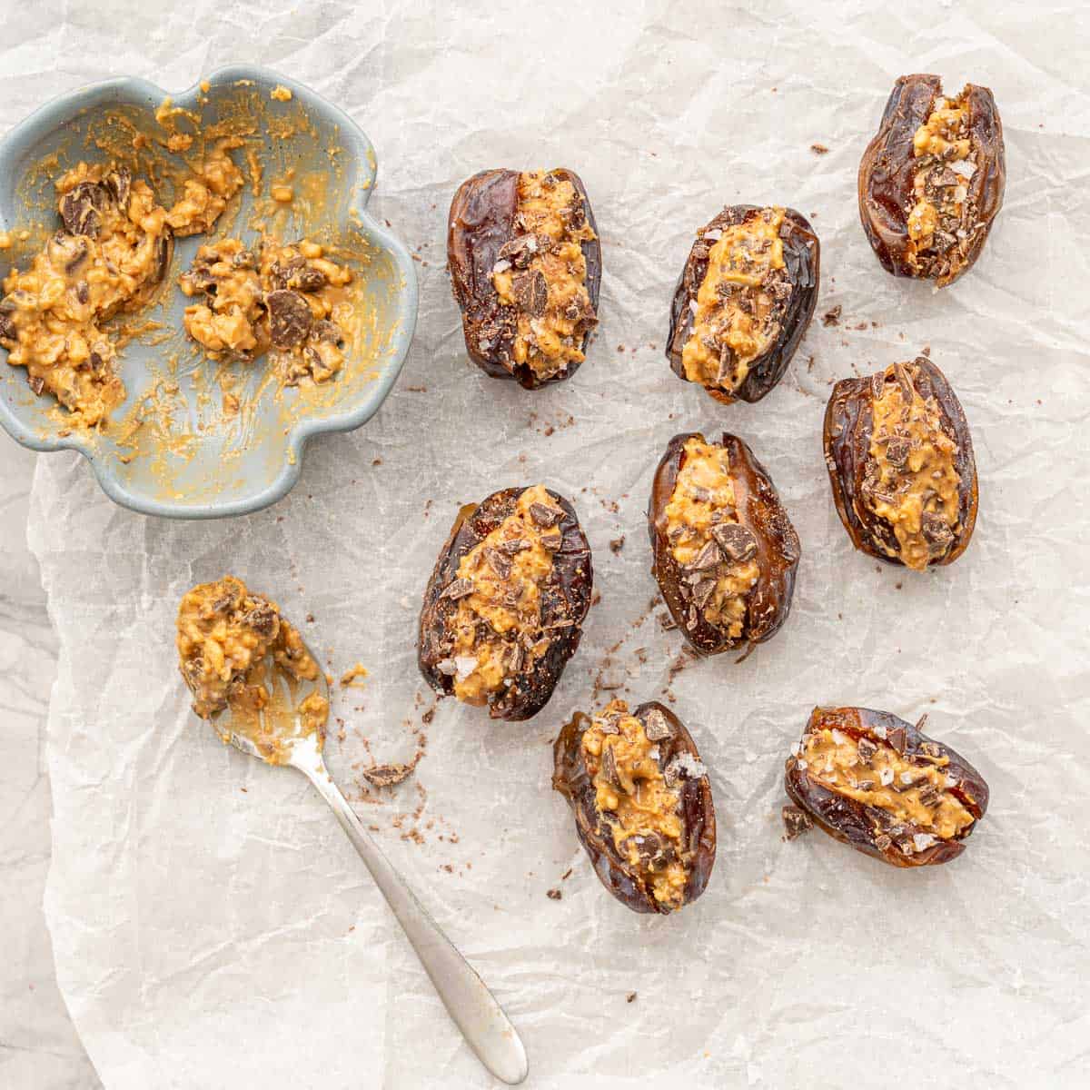 Peanut butter mixture stuffed inside cut open dates laid on baking paper with leftover mixture inside small ramekin and teaspoon.