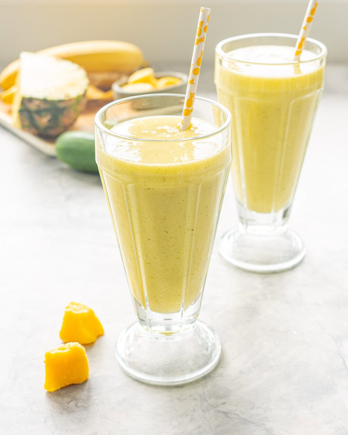 Two tall sundae glasses almost filled to the top with tropical smoothie inside and straw. Ingredients chopped and blurred in background.