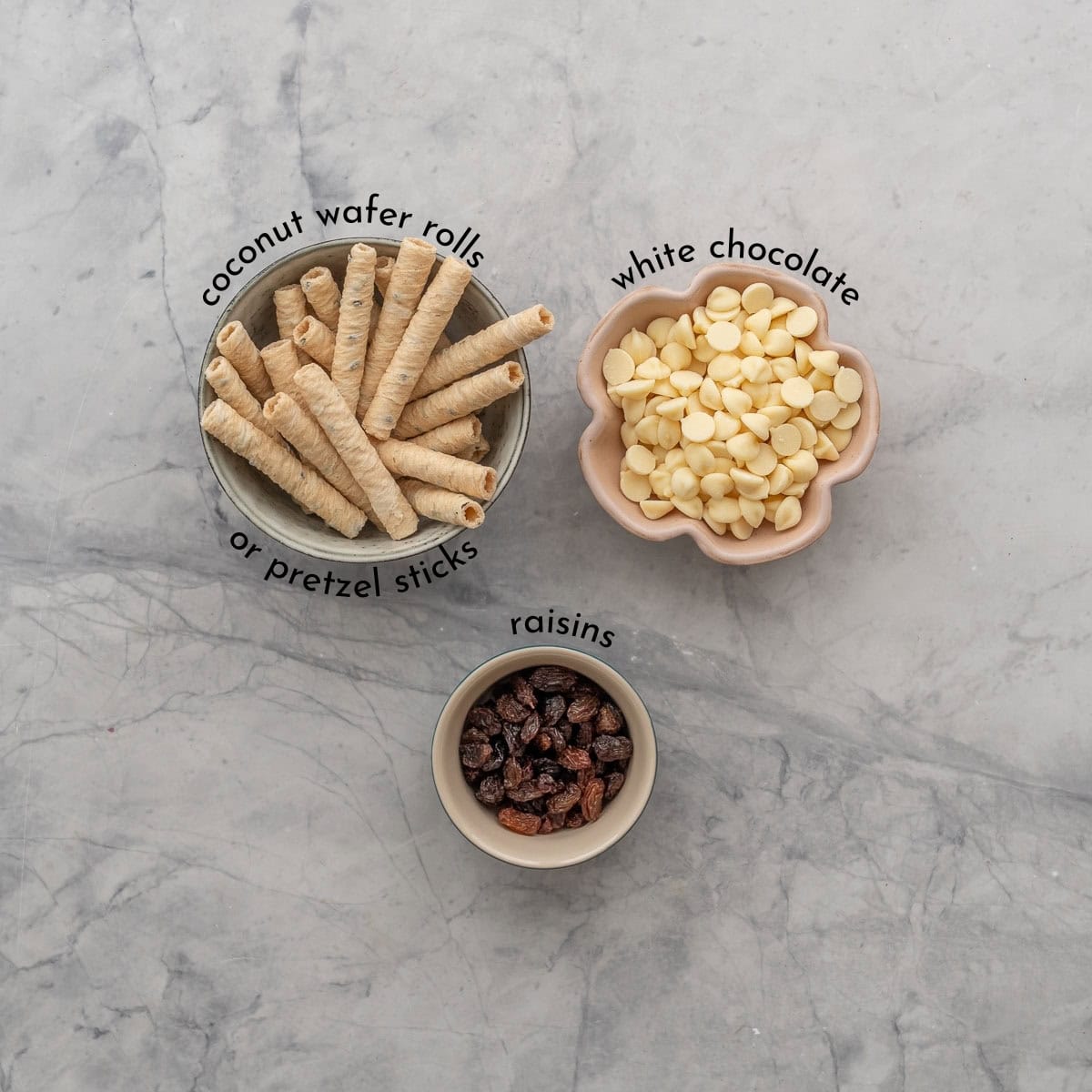 Ingredients for Spiderweb pretzles on benchtop with text overlay. Coconut wafer rolls, white chocolate chips and raisins.