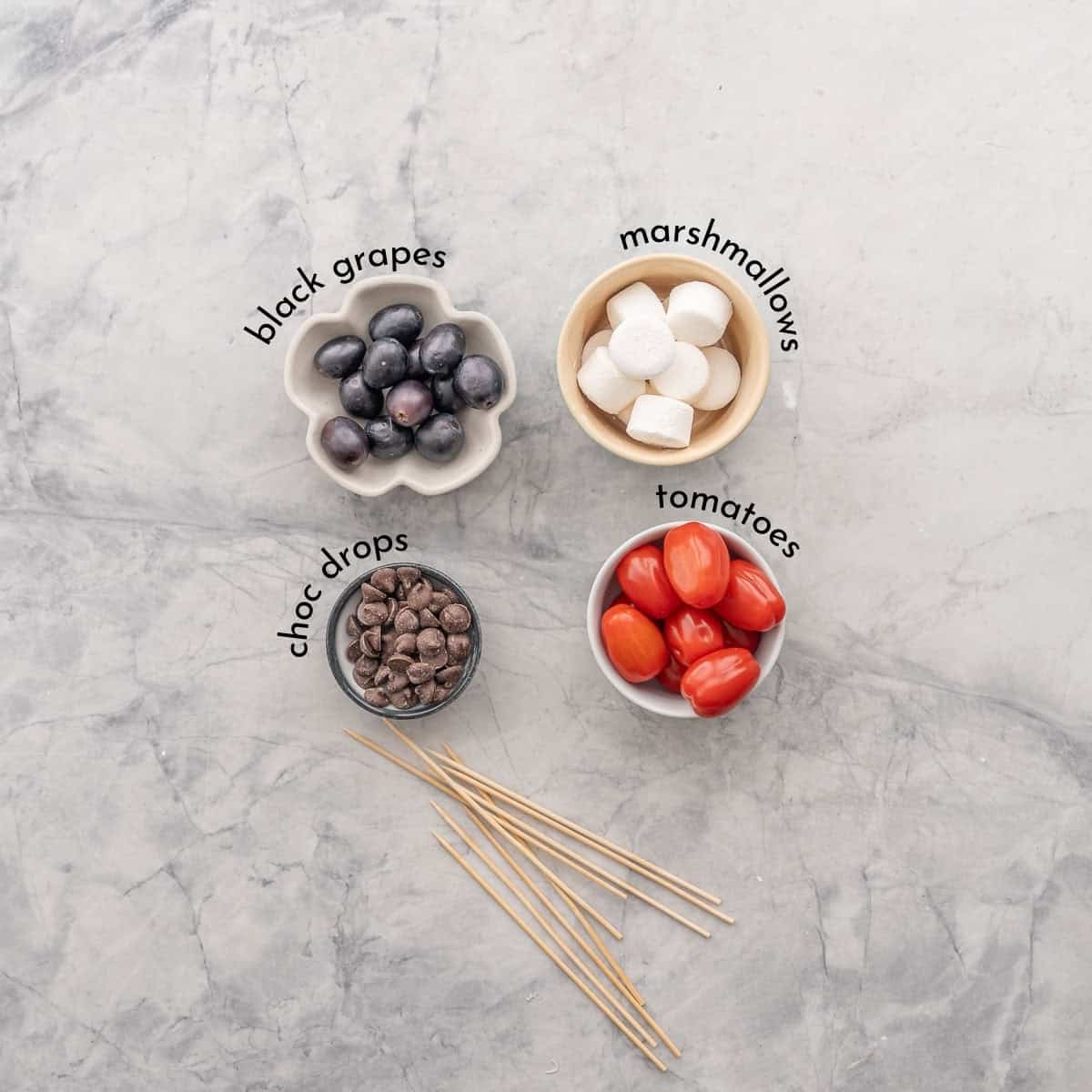 Ingredients for Fruit and Veggie Skewers on benchtop with text overlay. Black grapes, white marshmallows, chocolate drops, cherry tomatoes and skewers.