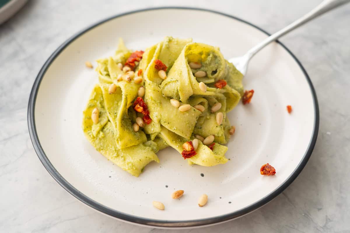 A portion of Avocado pasta served on a plate garnished with pine nuts and diced sun-dried tomato.