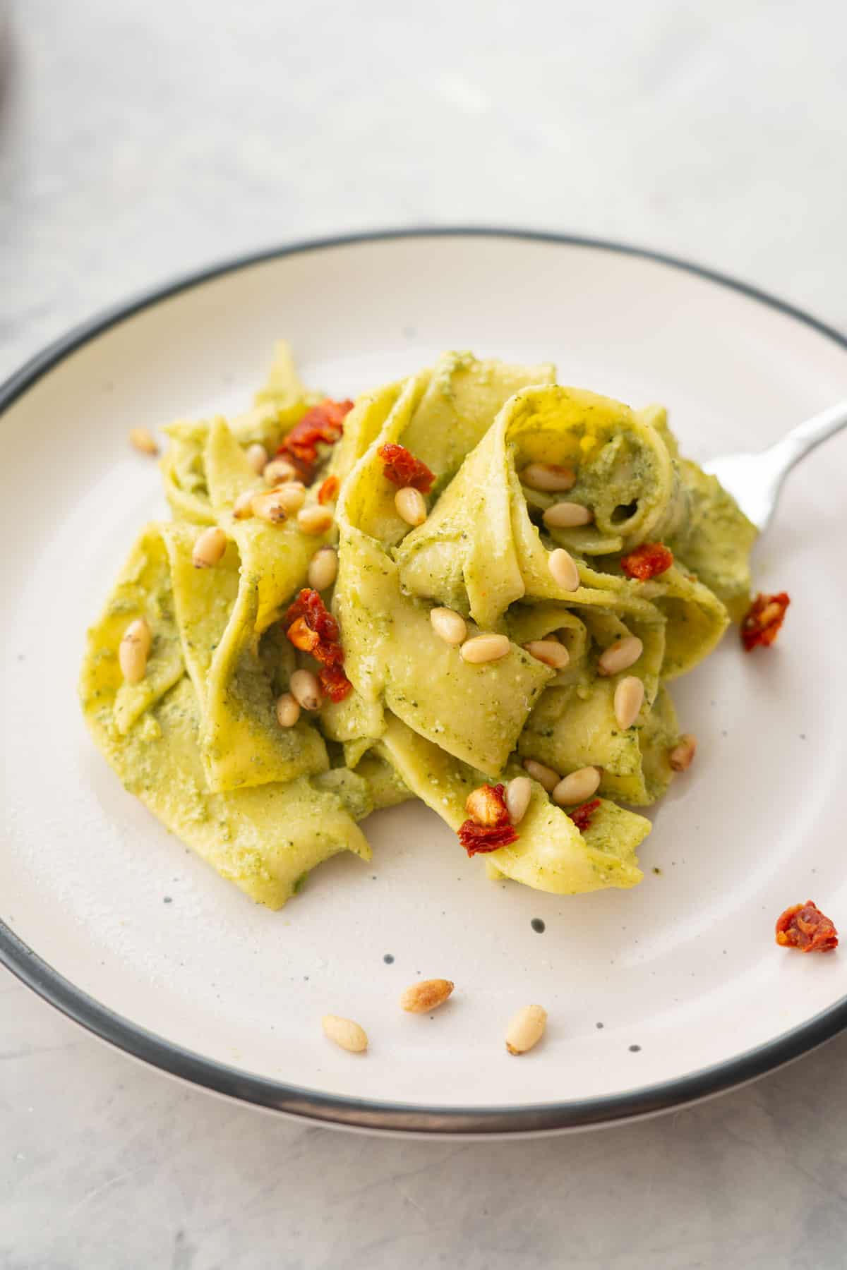 A portion of Avocado pasta served on a plate garnished with pine nuts and diced sun-dried tomato.