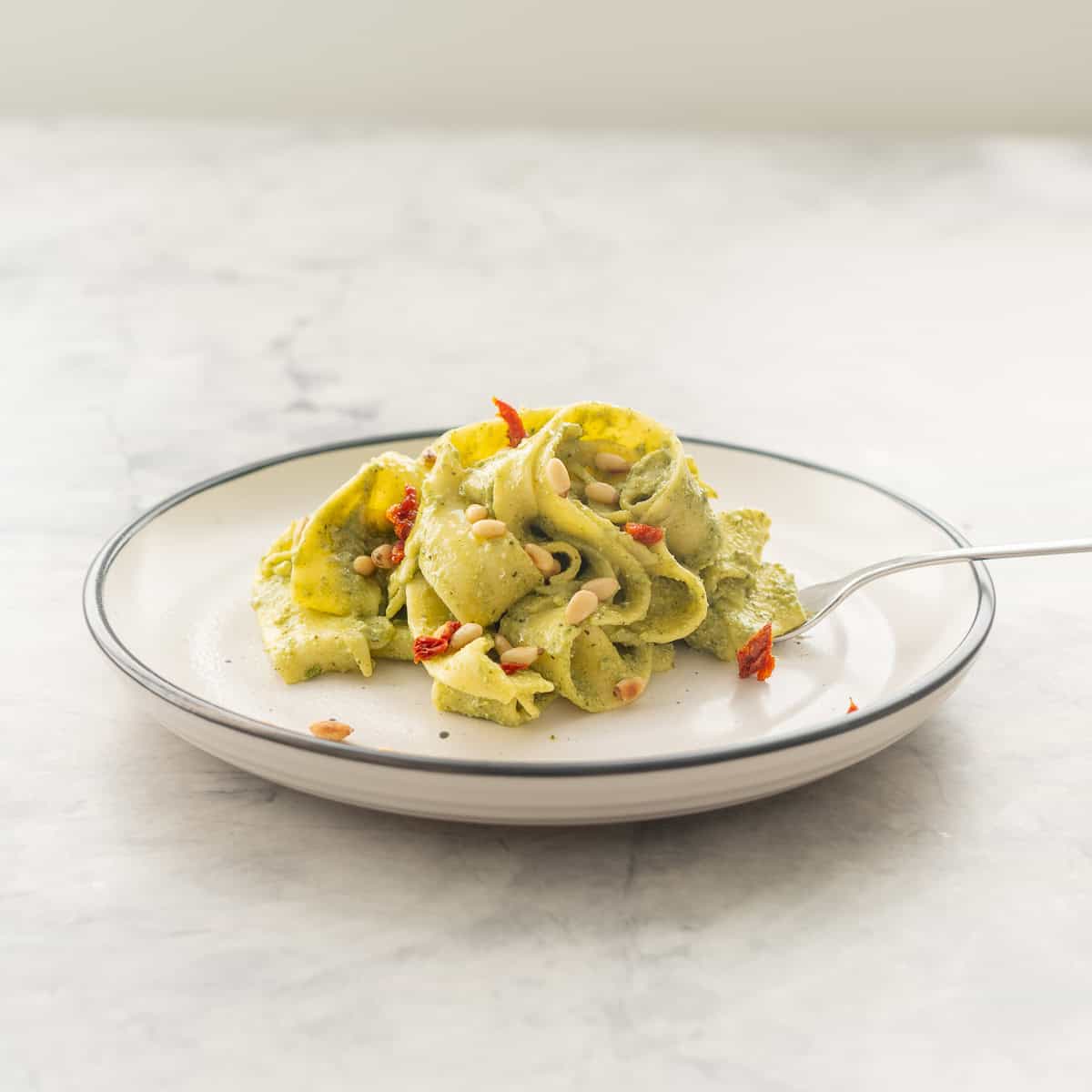 A portion of Avocado pasta served on a plate garnished with pine nuts and diced sun-dried tomato.