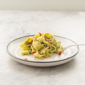 A portion of Avocado pasta served on a plate garnished with pine nuts and diced sun-dried tomato.