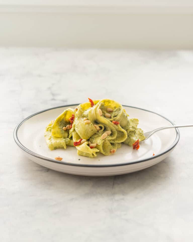 A portion of Avocado pasta served on a plate garnished with pine nuts and diced sun-dried tomato.