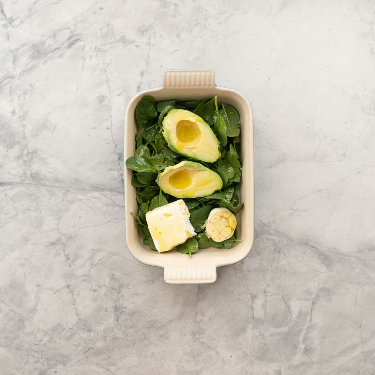 Fresh spinach leaves, peeled and pipped avocado cut in half, feta cheese and a garlic bolb with the head cut off, all drizzled with olive oil in a baking dish.