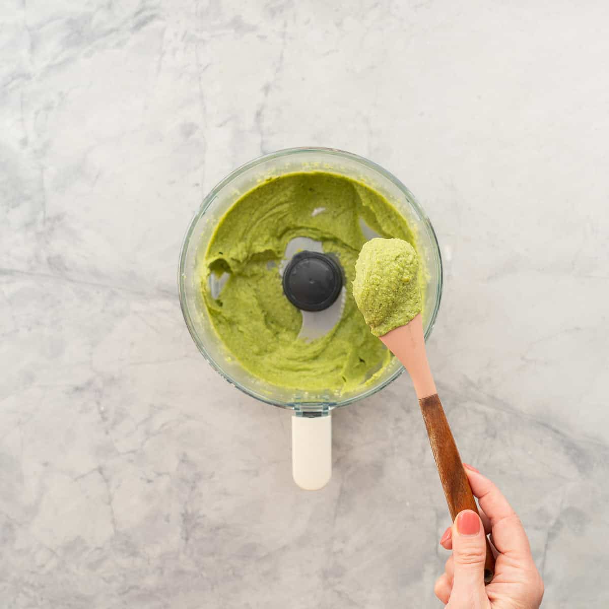 Food processor on bench top with smoothly blended avocado,feta,garlic and spinach being scooped onto a spatula.